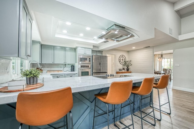 kitchen featuring stainless steel appliances, light stone countertops, a raised ceiling, kitchen peninsula, and light wood-type flooring