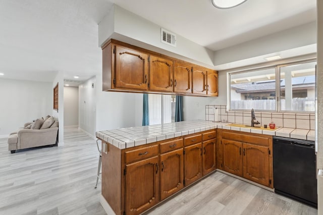 kitchen with sink, tile countertops, light wood-type flooring, black dishwasher, and kitchen peninsula