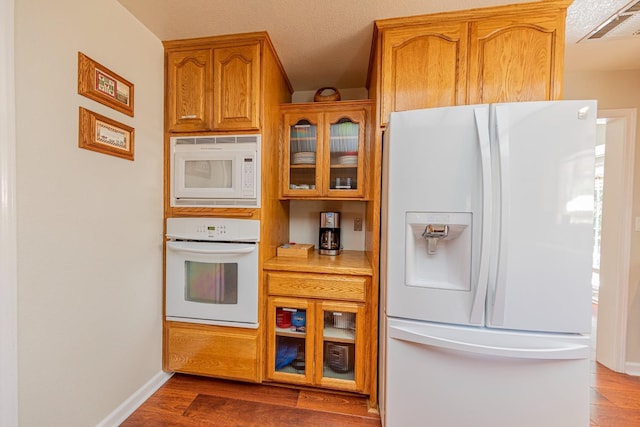 kitchen with white appliances, wood finished floors, light countertops, brown cabinetry, and glass insert cabinets