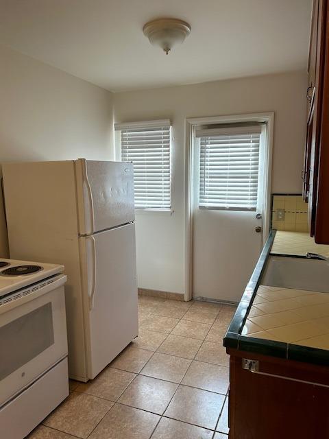 kitchen with dark brown cabinetry, white appliances, tile counters, a sink, and light tile patterned flooring