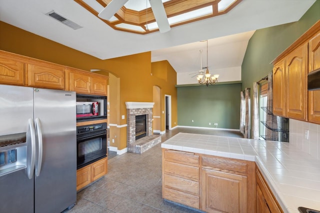 kitchen featuring black oven, stainless steel refrigerator with ice dispenser, tile counters, a brick fireplace, and decorative light fixtures