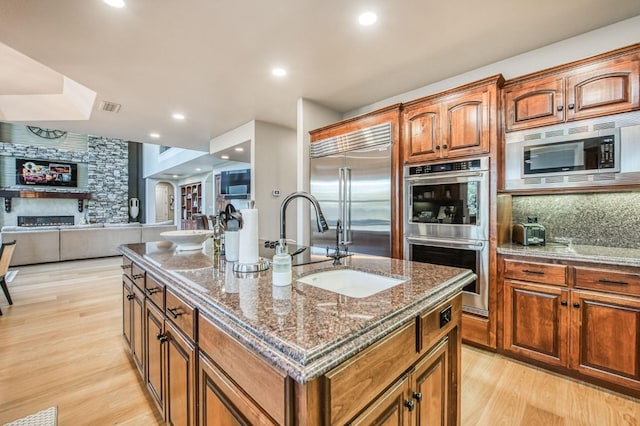 kitchen with dark stone countertops, built in appliances, light hardwood / wood-style floors, an island with sink, and decorative backsplash