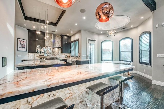 kitchen featuring wall chimney exhaust hood, hanging light fixtures, a raised ceiling, light stone countertops, and backsplash