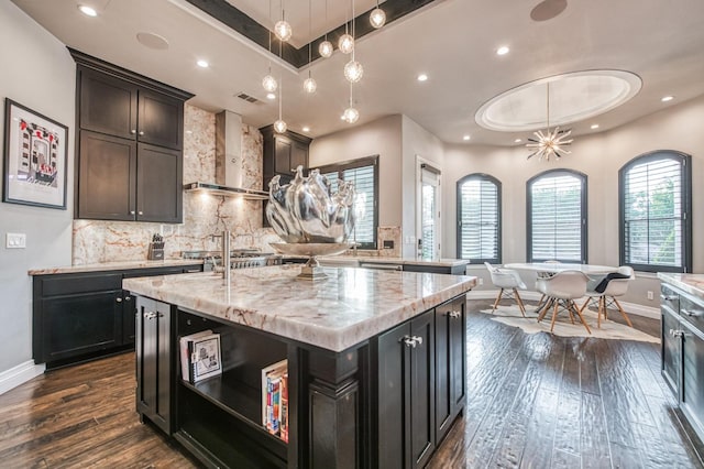 kitchen featuring pendant lighting, dark hardwood / wood-style floors, an island with sink, a raised ceiling, and wall chimney exhaust hood