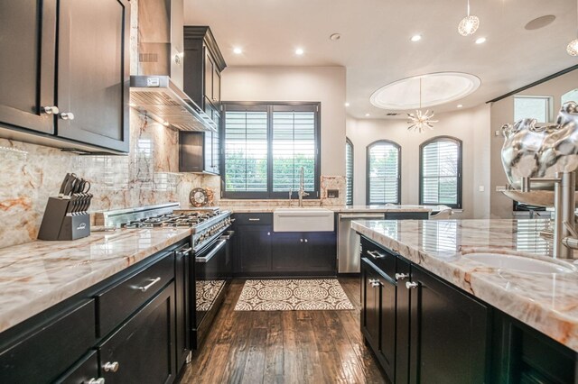 kitchen featuring wall chimney exhaust hood, light stone counters, decorative light fixtures, stainless steel appliances, and backsplash