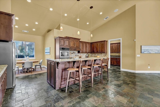 kitchen featuring appliances with stainless steel finishes, high vaulted ceiling, an island with sink, backsplash, and hanging light fixtures