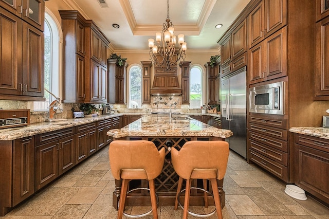 kitchen with built in appliances, a center island with sink, a kitchen breakfast bar, a raised ceiling, and pendant lighting