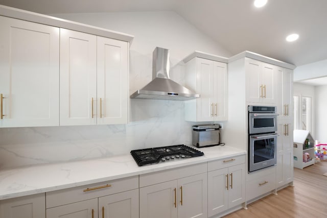 kitchen with vaulted ceiling, appliances with stainless steel finishes, white cabinets, light stone countertops, and wall chimney range hood