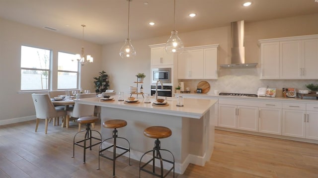 kitchen featuring pendant lighting, wall chimney range hood, white cabinetry, stainless steel microwave, and a center island with sink