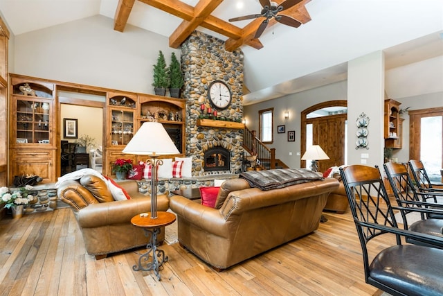 living room with ceiling fan, beam ceiling, high vaulted ceiling, a stone fireplace, and light wood-type flooring