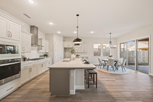 kitchen with an island with sink, white cabinetry, sink, stainless steel appliances, and wall chimney exhaust hood