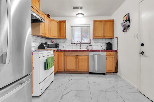 kitchen with tile counters, under cabinet range hood, appliances with stainless steel finishes, marble finish floor, and a sink