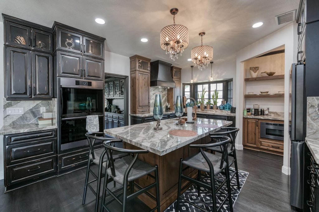 kitchen with hanging light fixtures, an island with sink, custom range hood, stainless steel appliances, and light stone countertops