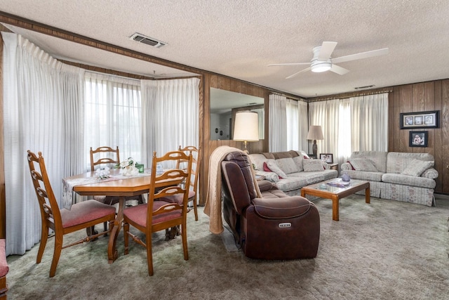 carpeted living area with ceiling fan, plenty of natural light, visible vents, and wooden walls