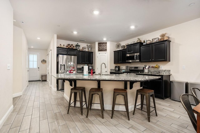 kitchen featuring stainless steel appliances, a kitchen island with sink, a breakfast bar area, and light stone counters