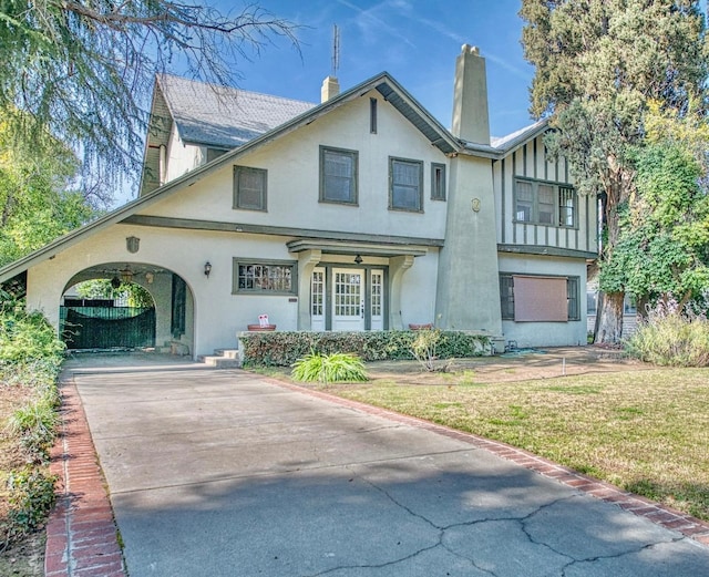 tudor-style house featuring driveway, a chimney, a front yard, and stucco siding