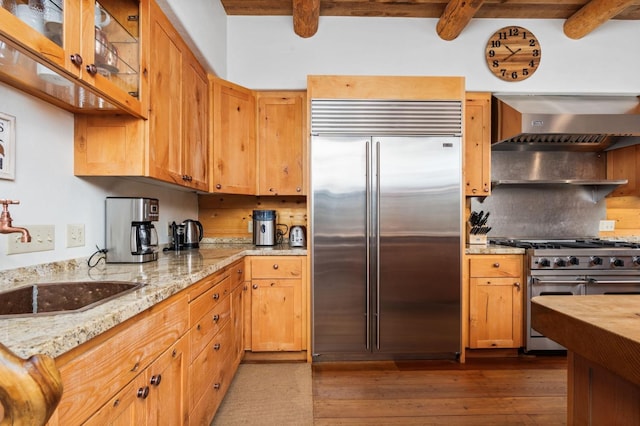 kitchen featuring beamed ceiling, sink, premium appliances, light stone counters, and wall chimney range hood