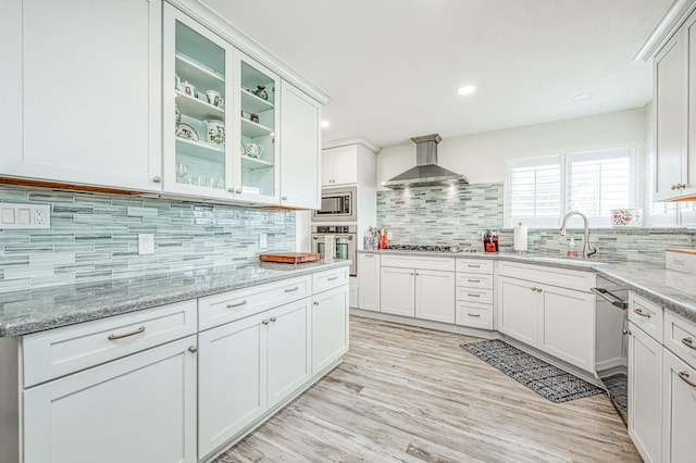 kitchen with wall chimney exhaust hood, sink, white cabinetry, light stone counters, and appliances with stainless steel finishes