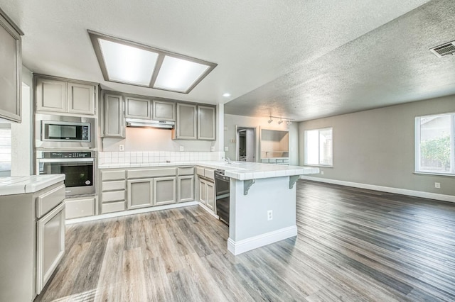 kitchen with gray cabinetry, under cabinet range hood, stainless steel appliances, a peninsula, and tile counters