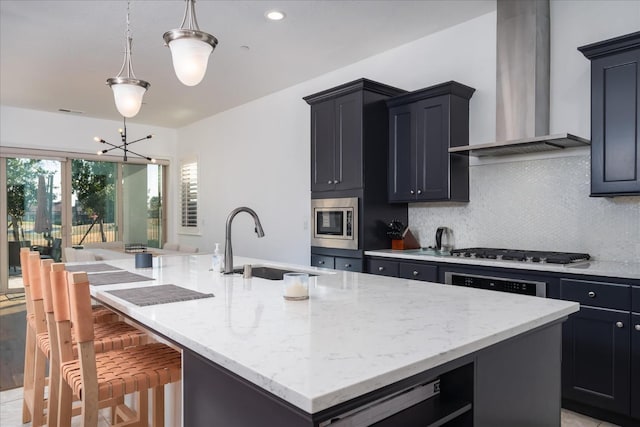 kitchen with appliances with stainless steel finishes, decorative light fixtures, sink, wall chimney range hood, and a center island with sink