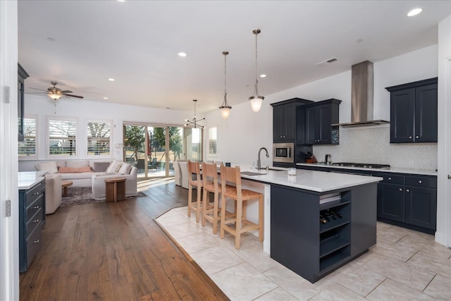 kitchen featuring appliances with stainless steel finishes, a kitchen bar, hanging light fixtures, a kitchen island with sink, and wall chimney exhaust hood