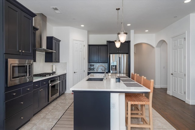 kitchen featuring a kitchen bar, sink, a center island with sink, stainless steel appliances, and wall chimney range hood