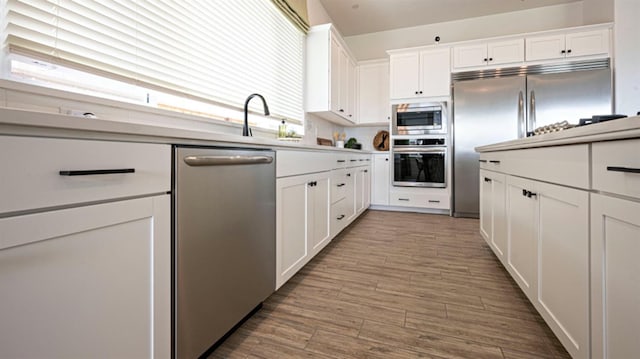 kitchen with a sink, built in appliances, light countertops, white cabinetry, and wood finished floors