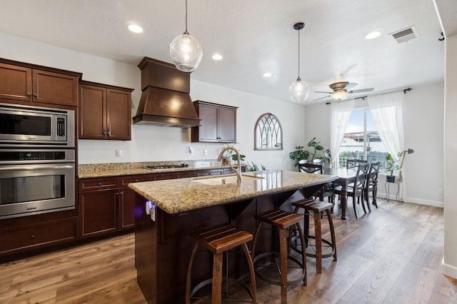 kitchen with premium range hood, sink, a center island with sink, appliances with stainless steel finishes, and light stone countertops