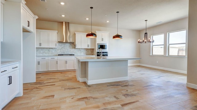 kitchen with an island with sink, white cabinetry, hanging light fixtures, stainless steel appliances, and wall chimney exhaust hood