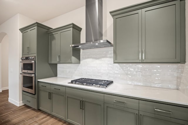 kitchen featuring wall chimney exhaust hood, light stone counters, hardwood / wood-style flooring, stainless steel appliances, and backsplash