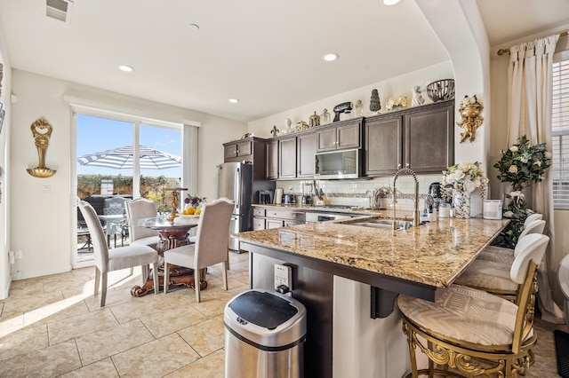 kitchen featuring a breakfast bar, stainless steel appliances, kitchen peninsula, dark brown cabinets, and light stone countertops