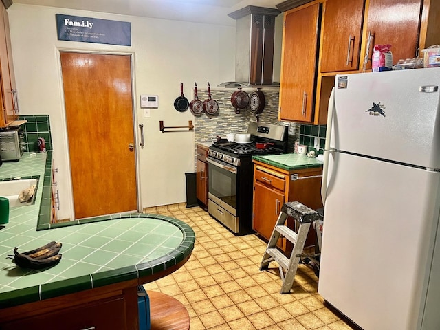 kitchen featuring stainless steel range with gas stovetop, wall chimney exhaust hood, tile counters, decorative backsplash, and white refrigerator