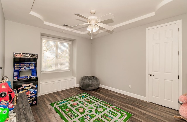 playroom with dark wood-type flooring, a raised ceiling, a ceiling fan, and baseboards