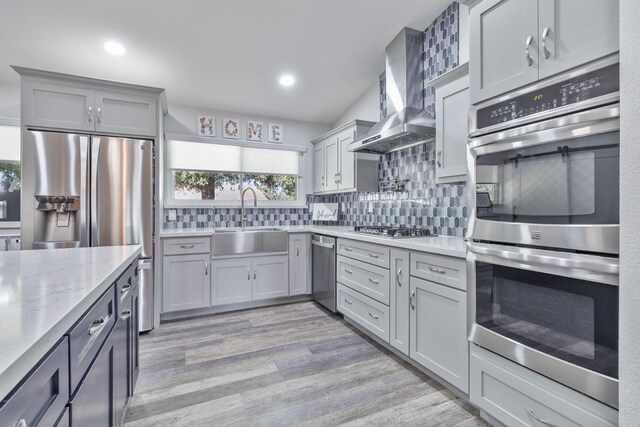 kitchen with tasteful backsplash, gray cabinets, appliances with stainless steel finishes, a sink, and wall chimney range hood