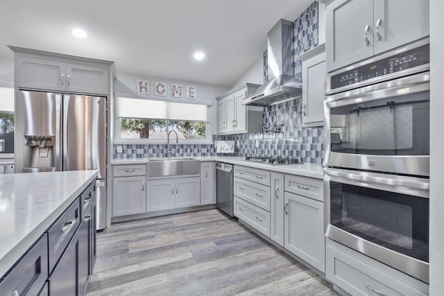 kitchen featuring stainless steel appliances, decorative backsplash, a sink, wall chimney range hood, and light wood-style flooring