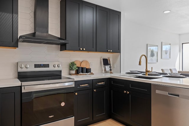 kitchen with light countertops, appliances with stainless steel finishes, a sink, dark cabinets, and wall chimney exhaust hood