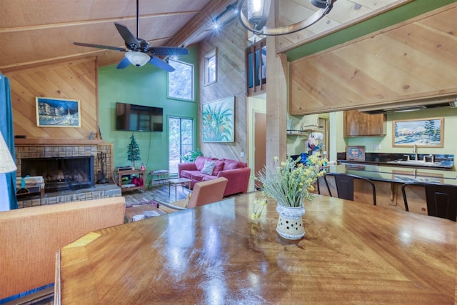 dining space with wood walls, ceiling fan, beam ceiling, a tiled fireplace, and high vaulted ceiling