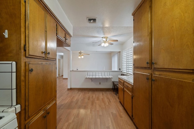 kitchen featuring a textured ceiling, a ceiling fan, light countertops, light wood-type flooring, and brown cabinetry