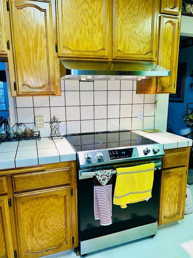 kitchen featuring stainless steel range with electric stovetop, range hood, tile countertops, and decorative backsplash