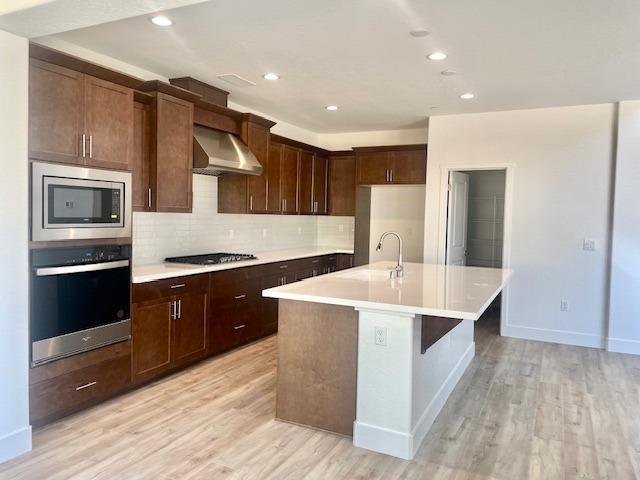 kitchen with appliances with stainless steel finishes, light countertops, a kitchen island with sink, and wall chimney exhaust hood