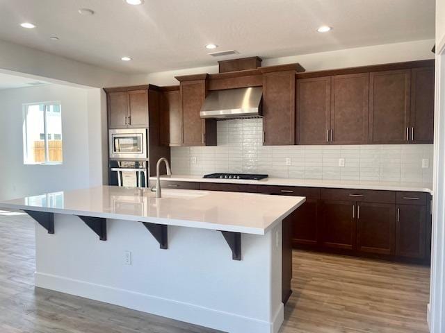 kitchen with a kitchen island with sink, a sink, light countertops, appliances with stainless steel finishes, and wall chimney exhaust hood