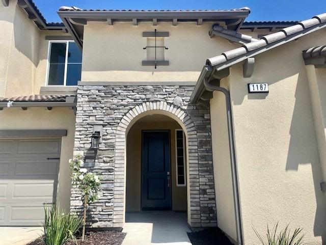 doorway to property featuring an attached garage, stone siding, a tile roof, and stucco siding