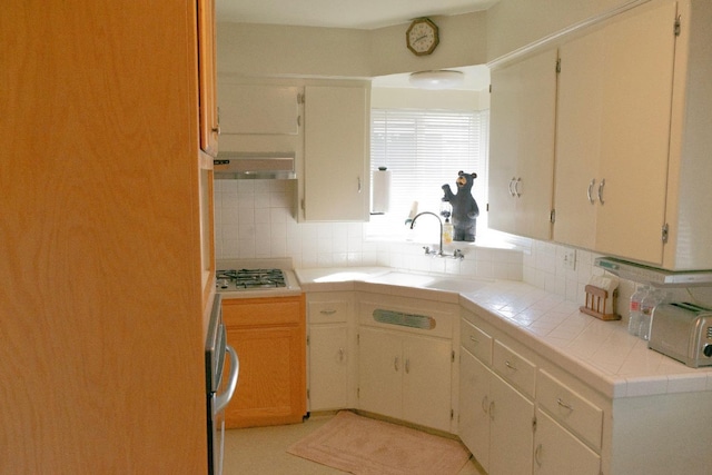 kitchen featuring cream cabinets, under cabinet range hood, stainless steel gas cooktop, tile counters, and tasteful backsplash