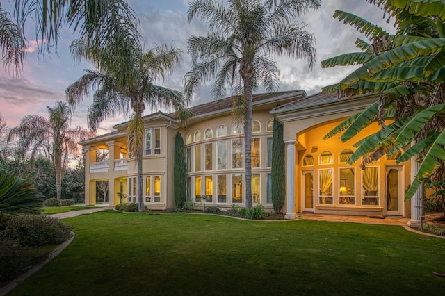 back of house at dusk with a lawn, a balcony, and stucco siding