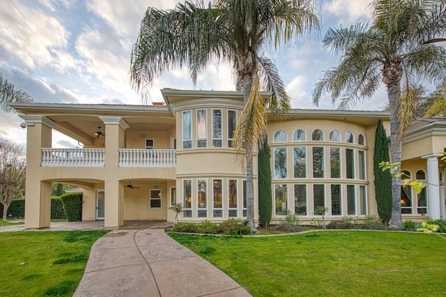 back of property with a yard, ceiling fan, a balcony, and stucco siding