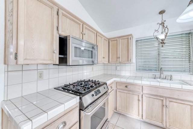 kitchen with tile countertops, hanging light fixtures, stainless steel appliances, light brown cabinets, and a sink