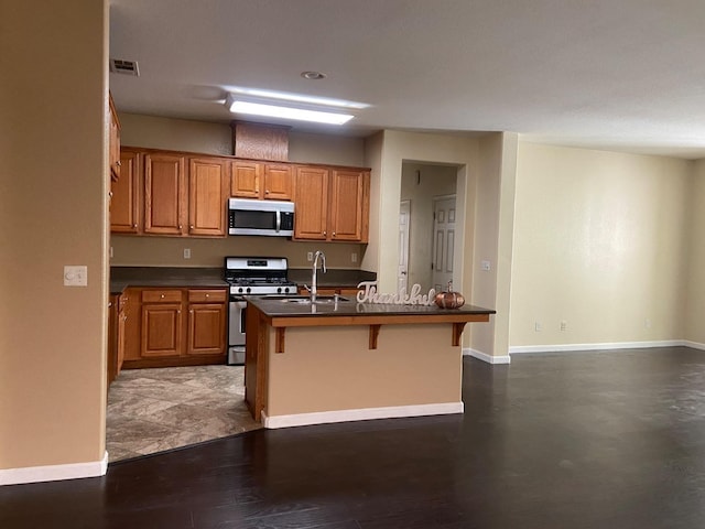 kitchen featuring a center island with sink, brown cabinetry, dark countertops, appliances with stainless steel finishes, and a kitchen bar