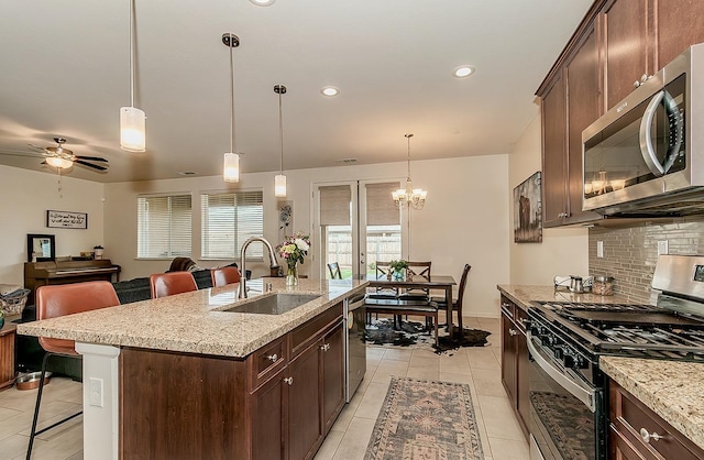 kitchen featuring stainless steel appliances, a sink, hanging light fixtures, light stone countertops, and a center island with sink