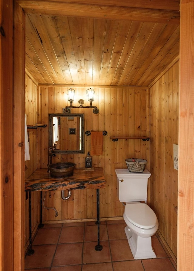 half bathroom featuring wooden ceiling, toilet, wood walls, a sink, and tile patterned floors