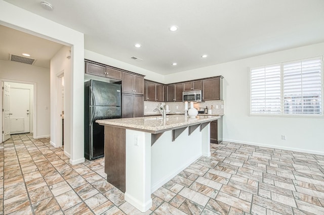 kitchen featuring tasteful backsplash, visible vents, stainless steel microwave, freestanding refrigerator, and a kitchen island with sink